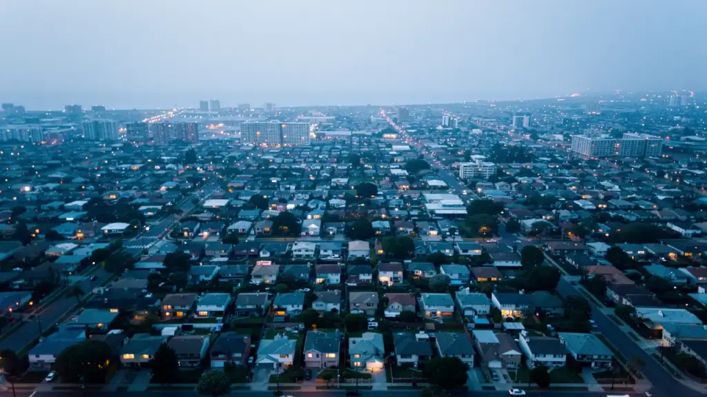 A coastal city grid at dusk with the first warm window lights coming on across the neighborhood.